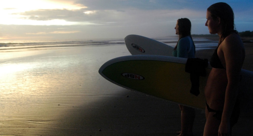 Two people hold surfboards while standing on a beach reflecting the gentle colors of the sky.